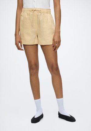 Persoon die beige shorts met trekkoord, witte sportsokken en zwarte platte schoenen draagt, staat tegen een effen lichte achtergrond.