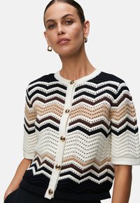 Hæklede cardigan med zigzag mønster i creme, brun og marineblå, med seks guldknapper og korte ærmer, lavet af et tekstureret strik.
