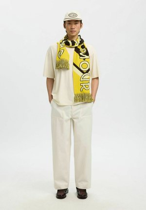 Mann iført kremfarget antrekk med gult og svart mønstret skjerf, kremfarget lue og mørkebrune sko, stående med hendene i lommene.