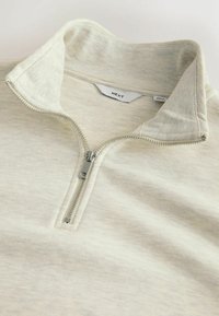 Light beige zip-up pullover with a mock collar, made of textured fabric, featuring a metallic zipper and label tag inside the collar.