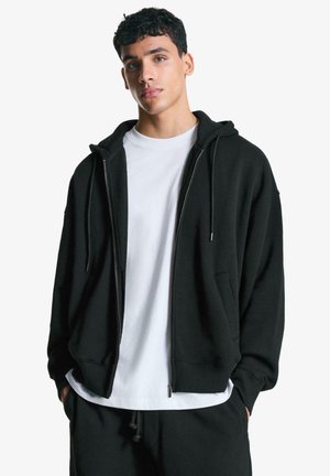 Black zip-up hoodie with a drawstring hood, ribbed cuffs, and front pockets, layered over a white t-shirt. Soft fabric, relaxed fit.