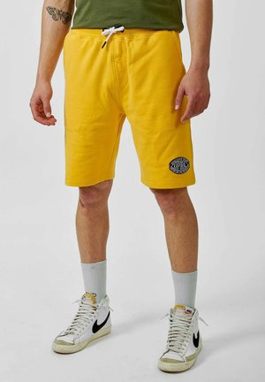 Shorts en coton jaune avec une taille élastique et un cordon de serrage, comportant un patch logo circulaire noir sur le côté gauche. Portés avec des baskets blanches.