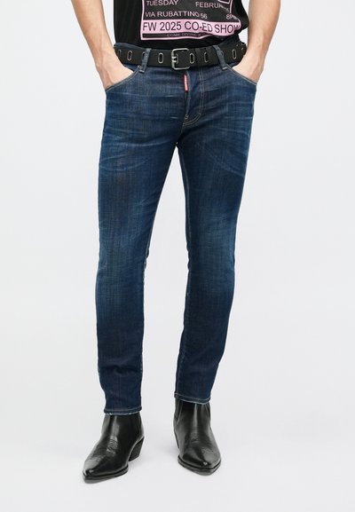 Jeans azul oscuro con un corte ajustado, que presentan un ligero desgaste, diseño clásico de cinco bolsillos, combinados con botas de vaquero negras y un cinturón.