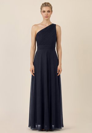 Frau steht nach vorne blickend und trägt ein ärmelloses, marineblaues, ein-Schulter-Abendkleid bodenlang mit plissiertem Oberteil.