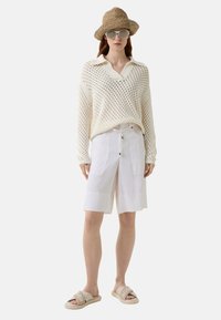 Lichte gebreide trui met V-hals, witte knielange shorts en beige slidesandalen met opdruk. Ronde zonnebril en strooien hoed.