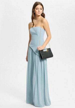 Jeune femme vêtue d'une robe bustier bleu clair tenant un sac à main noir texturé avec une chaîne dorée, sur fond blanc.