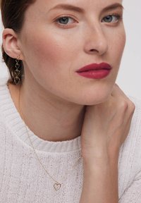 Złote kolczyki w kształcie serca oraz pasujący złoty naszyjnik w kształcie serca na kobiecie noszącej białą sweter z fakturowanym wzorem, widoczne odcienie skóry i cechy twarzy.