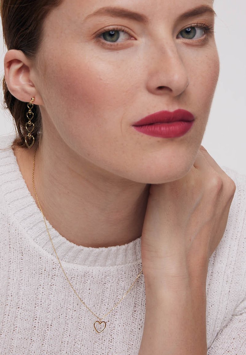Złote kolczyki w kształcie serca oraz pasujący złoty naszyjnik w kształcie serca na kobiecie noszącej białą sweter z fakturowanym wzorem, widoczne odcienie skóry i cechy twarzy.