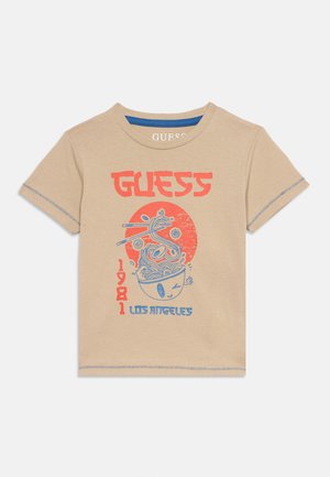 T-shirt beige à manches courtes avec texte rouge "GUESS", graphisme bleu et rouge représentant un bol de nouilles clignant de l'œil avec des baguettes, texte "1981" et "LOS ANGELES".