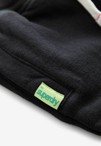 Čierna mikina z látky s hladkou textúrou, so zelenou vyšitou etikettou, ktorá na dolnom okraji znie "by superdry".