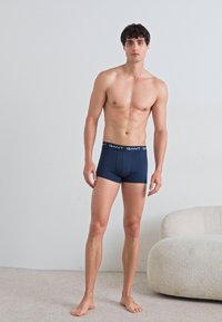 Marineblaue Boxershorts mit glatter Oberfläche, ausgestattet mit einem kontrastierenden schwarzen Bund, auf dem der Markenname "GANT" in wiederholtem weißen Schriftzug zu sehen ist.