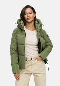 Groene pufferjack met een capuchon, voorzien van gestikte patronen en een rits, gedragen over een lichtbeige top en crèmekleurige broek.