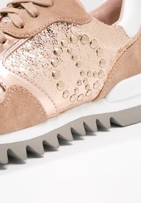 Gros plan d'une basket en or rose avec un panneau latéral métallique texturé, un motif de clous circulaires, des accents en daim beige et une semelle grise en dents de scie.