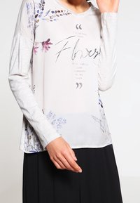 Maglietta leggera a maniche lunghe di un grigio chiaro con stampe floreali in viola e rosa. Presenta una citazione testuale centrata sul davanti. Tessuto morbido.