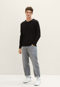 Pulover ribbed negru cu mâneci lungi și decolteu în V, asortat cu blugi deschiși la culoare cu manșete răsucite și teneși albi, pe un fundal neutru.
