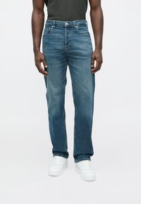 Calças de ganga azul com corte reto, design de cinco bolsos, com desbotamento subtil e fecho de botão, usadas com ténis brancos.