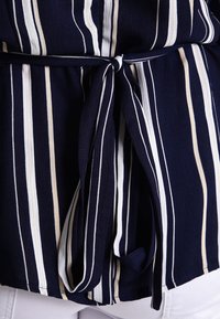 Tessuto blu navy con righe verticali bianche e beige annodato in vita sopra pantaloni bianchi.