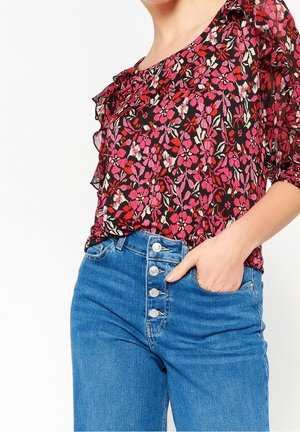 Donna che indossa una camicetta floreale rossa e rosa con volant e jeans blu a vita alta con quattro bottoni, mano in tasca.