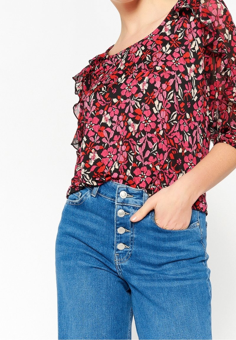 Femme portant un chemisier floral rouge et rose avec des volants et un jean bleu taille haute avec quatre boutons, main dans la poche.