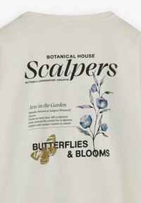 Camiseta de algodón color crema con estampados florales en azul y dorado, con un texto en negrita que detalla "Scalpers" y "Mariposas y Flores."