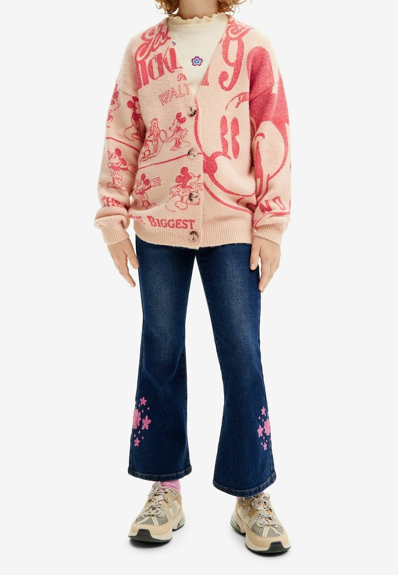 Enfant portant un gilet Mickey Mouse rose et rouge, un col roulé crème, un jean évasé bleu foncé avec des motifs de fleurs roses et des baskets beiges.