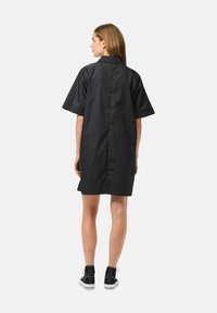 Abito camicia nero a maniche corte con taglio dritto, cucitura posteriore e orlo leggermente arrotondato. Indossato con stivali neri alla caviglia.