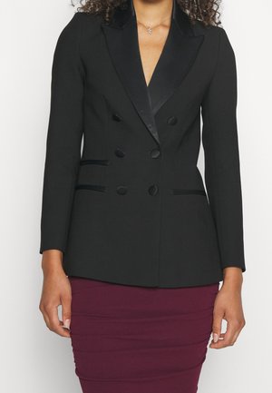 Woman wearing a fitted black double-breasted blazer with satin lapels and a burgundy ruched skirt, standing against a plain background.