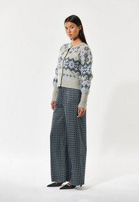 Grå strikket cardigan med blå blomster mønstre, parret med brede bukser med mønster i mørke nuancer, og sorte spidse sko.