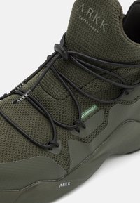 Zapatilla verde impermeable con parte superior de malla texturizada, cordones flexibles y suela de goma estructurada. Presenta el logo "ARKK" y un diseño que brinda soporte.