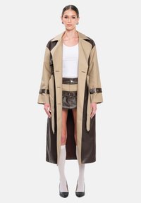 Trench-coat beige et marron avec larges revers, taille ceinturée et manches longues ; superposé à un haut blanc et une jupe en cuir marron.