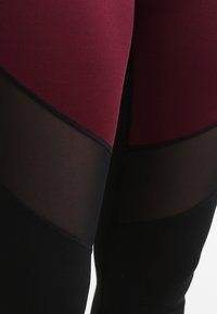 Lähikuva legginsseistä, joissa on burgundin värinen kangas, musta alaosa ja läpinäkyvät mustat verkko-osat reidissä.