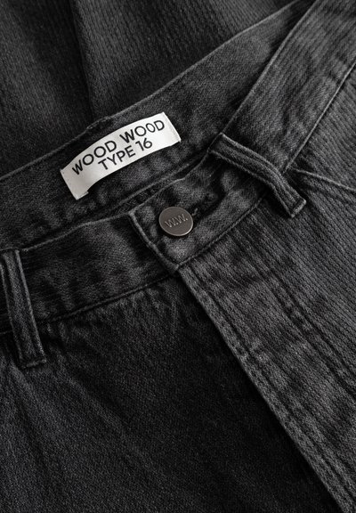 Fekete farmernadrág egyenes derékrésszel, márkajelzéssel ellátott címkével, fém gombos záródással és finom textúrával. A címkén az "WOOD WOOD TYPE 16" felirat olvasható.