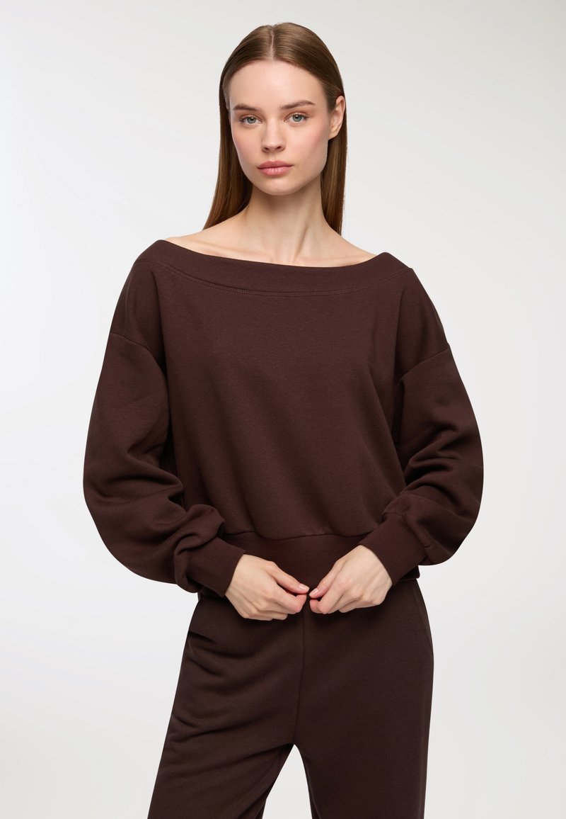 Felpa marrone con spalla scoperta, maniche lunghe, vita aderente e texture morbida. Il modello posa con le mani sui fianchi, indossando pantaloni coordinati.