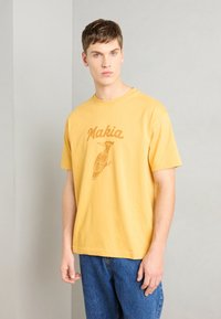 Gul bomulls-T-skjorte med "Makia"-tekst og grafikk av en brun fugl, med korte ermer og en avslappet passform. Kombinert med blå jeans.