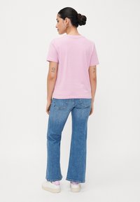 Roza bombažna majica s kratkimi rokavi, prikazana s hrbtne strani. Ujemajo se z modrimi visokimi flare jeans hlačami in belimi čevlji z detajli.