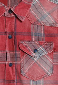 Červená kostkovaná košile s límcem na knoflíky, náprsní kapsou s klopou a tmavě modrými knoflíky. Vzor zahrnuje protínající se linie v různých odstínech.