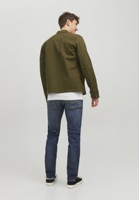 Giacca di cotone verde oliva con taglio dritto, abbinata a jeans blu e sneakers nere, che mostra un design semplice e casual.