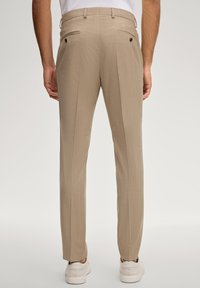 Mann in beigefarbenen, maßgeschneiderten Hosen und weißen Sneakers, von hinten vor einem einfarbigen hellen Hintergrund gezeigt.