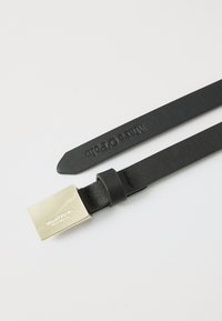 Ceinture en cuir noir avec une boucle rectangulaire en métal doré et un logo en relief. La sangle est texturée et lisse, avec des extrémités effilées.