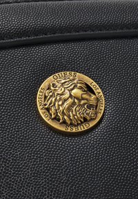 Surface en cuir noir texturé avec un médaillon doré rond présentant un lion rugissant et le texte "GUESS LOS ANGELES" autour du bord.