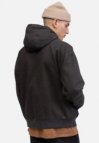 Black hooded jacket made of sturdy fabric, featuring a ribbed hem and cuffs, with an underlayer of a tan shirt visible underneath.