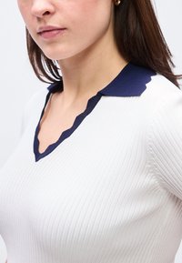 Γυναίκα που φοράει λευκή ριμπ μπλούζα με σκούρο μπλε γιακά σε σχήμα κυματιστού άκρου, δείχνοντας τον λαιμό της και μέρος από το πρόσωπο και τα μαλλιά της.