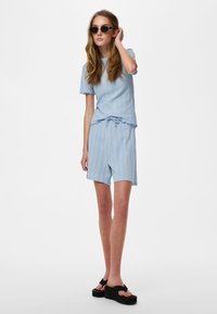 Jauna sieviete valkā gaiši zilu adījumu topu un šortus, melnas platformas sandales un apaļas melnas saulesbrilles uz vienkrāsaina balta fona.