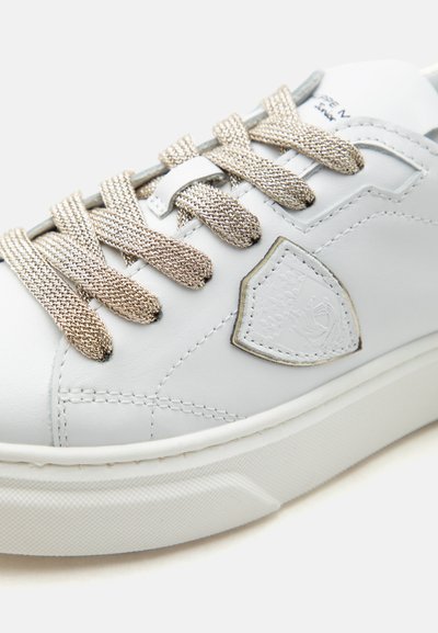 Basket en cuir blanc avec des lacets beige texturés, des détails cousus et un patch en forme de bouclier sur le côté près de la semelle.