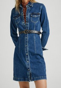 Jeansskjortklänning med långa ärmar, knäppning fram, två bröstfickor och ett svart mönstrat bälte i midjan, med en figurnära design.