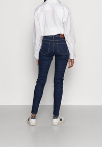 Smala mörkblå denimjeans med bakfickor, bärs med en vit skjorta med krage och vita sneakers med svarta detaljer.