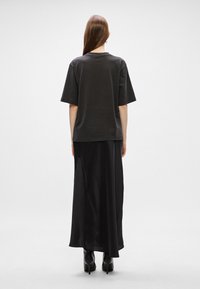 Μαύρη κοντομάνικη μπλούζα σε χαλαρή γραμμή, συνδυασμένη με μια μακριά μαύρη σατέν φούστα και μπότες με ψηλό τακούνι, βλέπεται από πίσω.