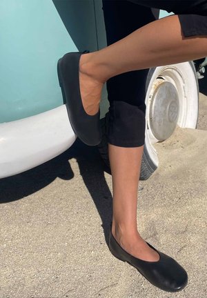 Benedenbenen met zwarte ballerina's en zwarte broek, rustend op de bumper en band van een lichtblauw vintage voertuig op een zandige ondergrond.