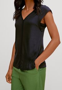 Top nero a maniche corte con scollo a V in tessuto morbido ed elastico, caratterizzato da una cucitura frontale e abbinato a pantaloni verdi.