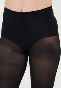 Primo piano di una persona che indossa collant neri opachi a vita alta con gambe trasparenti su uno sfondo neutro.